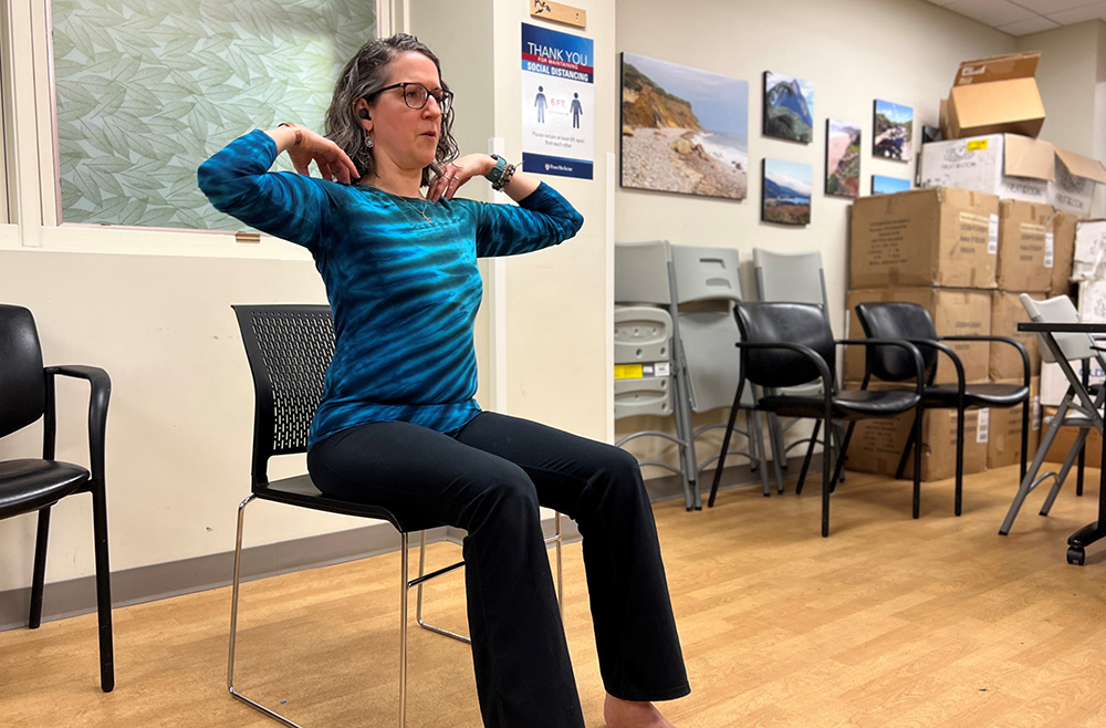 HUP secretary and volunteer yoga teacher Judy Glass sits in a chair with her hands touching her shoulders. 